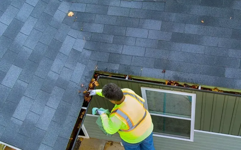 A worker in a hi-vis vest scooping leaves out of a gutter from a ladder